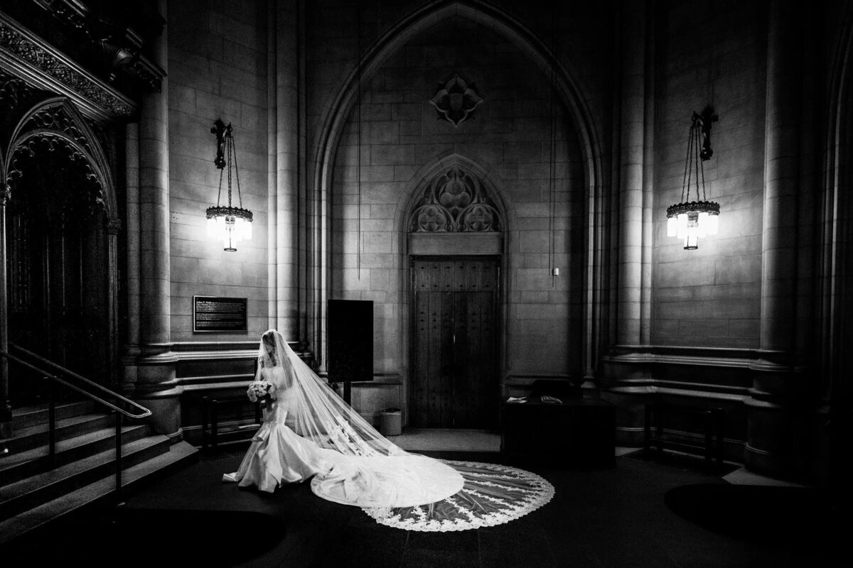 Black and white image of a bride in an elegant gown with a long veil, standing in the ornate doorway of a grand building