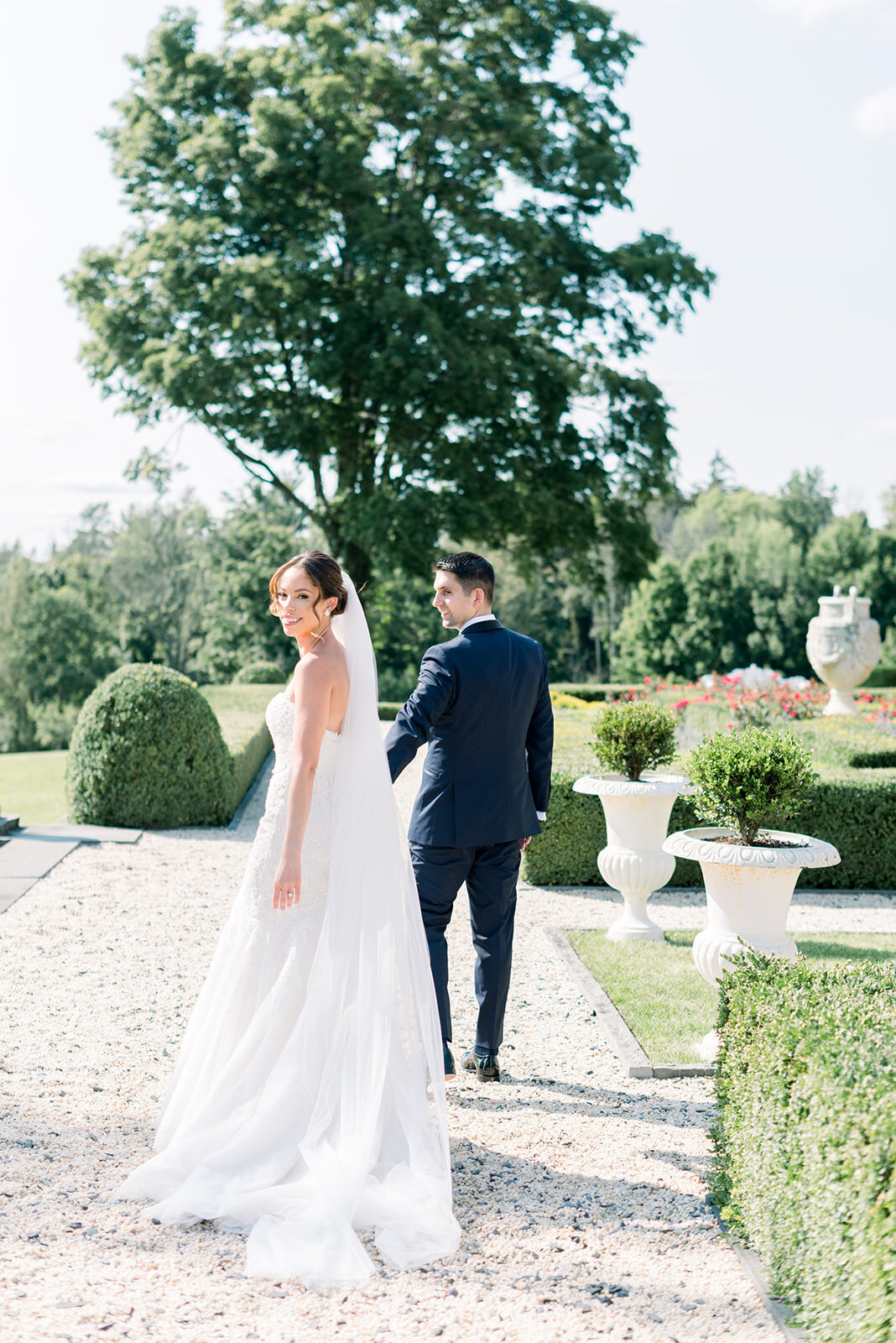 A bride smiles over her shoulder while walking hand in hand through a large garden with her groom