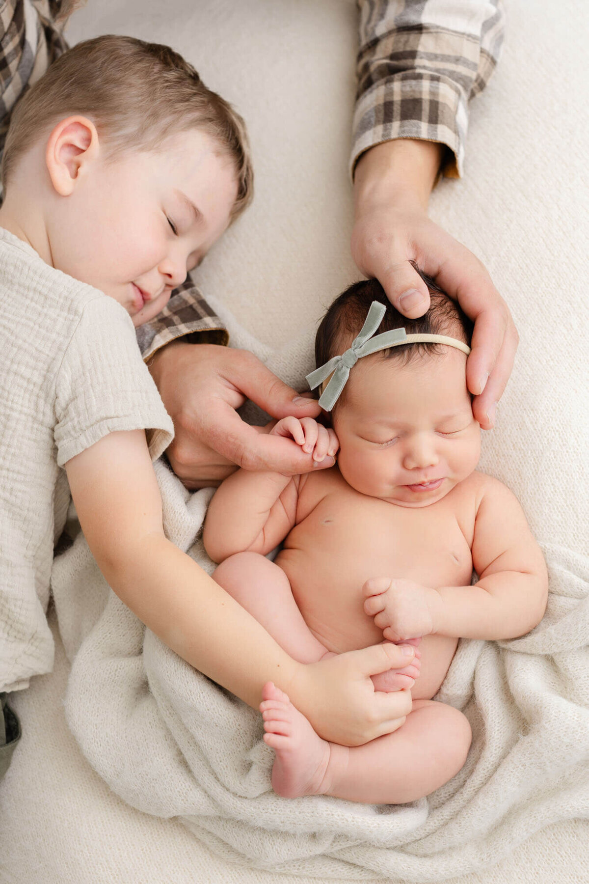 Dad holding baby girl while she is sleeping on beige blanket. Big brother is holding her foot and laying his head on dad's arm with his eyes closed. Light neutral tones and colors - PDX newborn photography