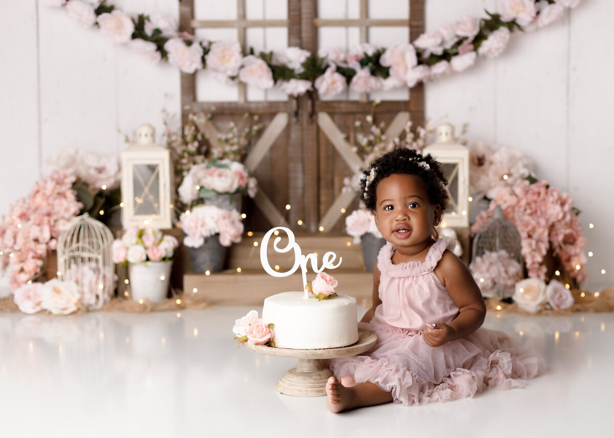 Farmhouse theme cake smash in Wellington and Boca Raton Florida. Baby girl is wearing a soft pink tutu dress sitting beside a white cake. In the background is a farm inspired backdrop with barn doors and floral garland.  Surrounding the backdrop are soft pink flowers with birdcages and lanterns