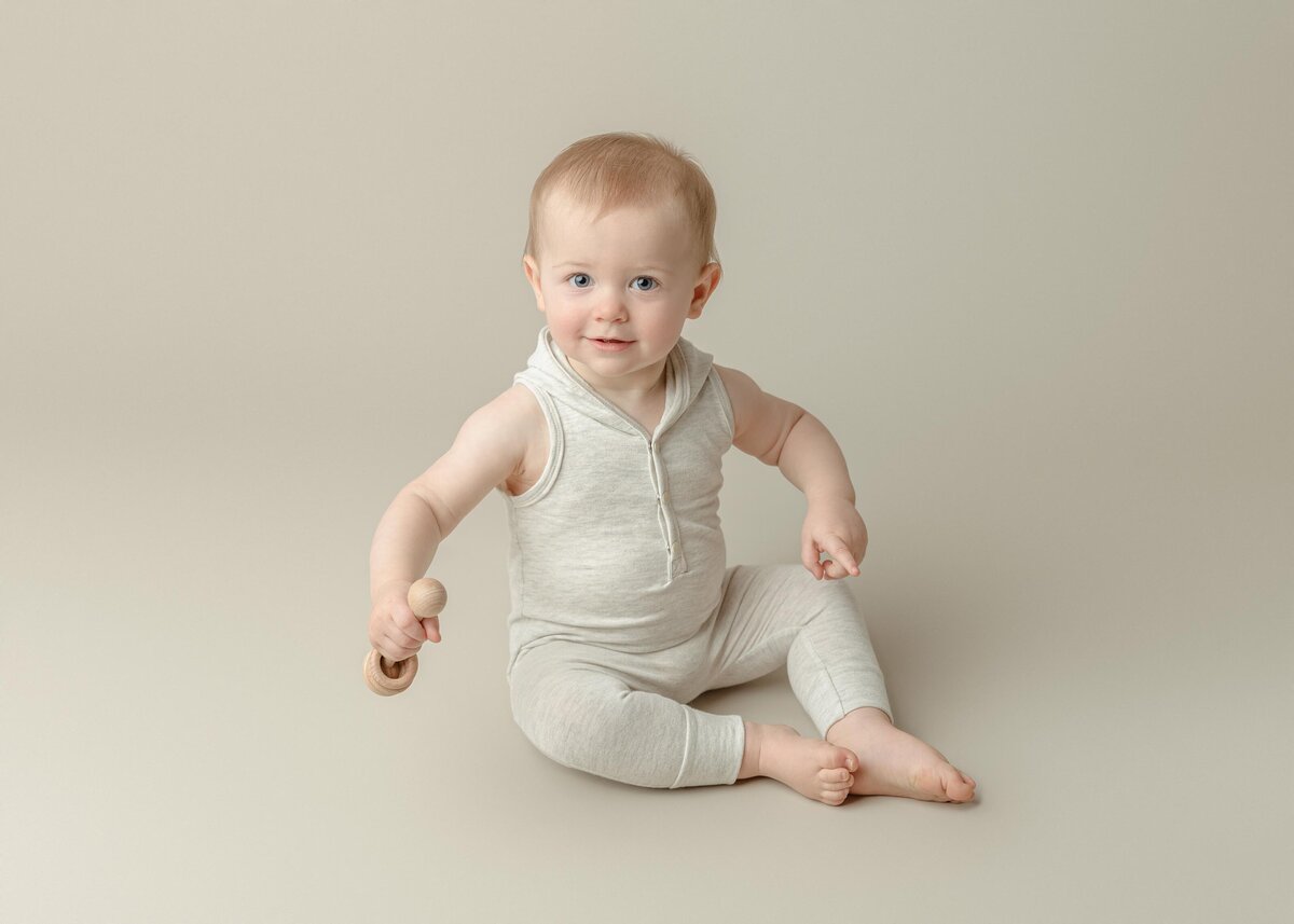 One. Year Old Boy In Oatmeal Outfit Sitting On Tan Backdrop Holding Rattle