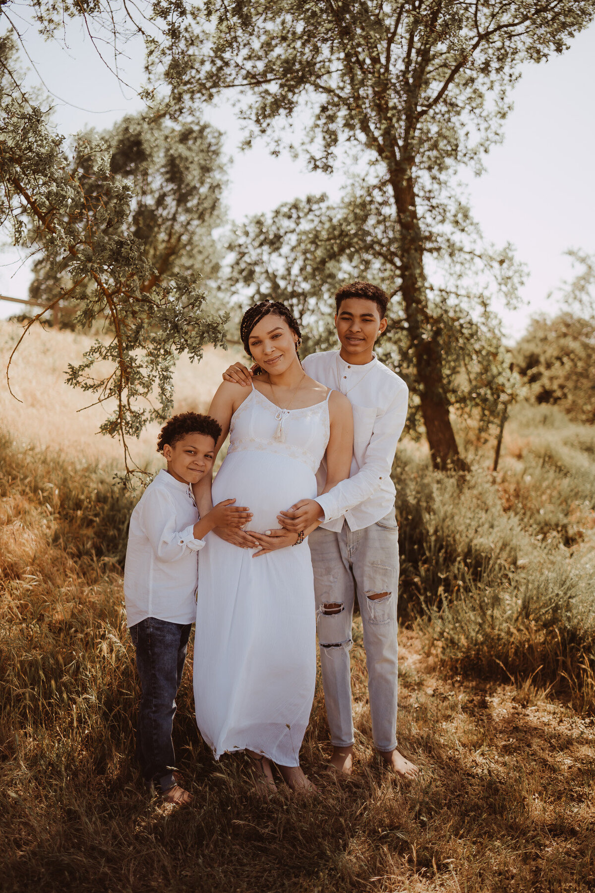 a woman expecting a baby in a white dress, woman holding her belly, her two sons hugging her from the side, hands on moms belly