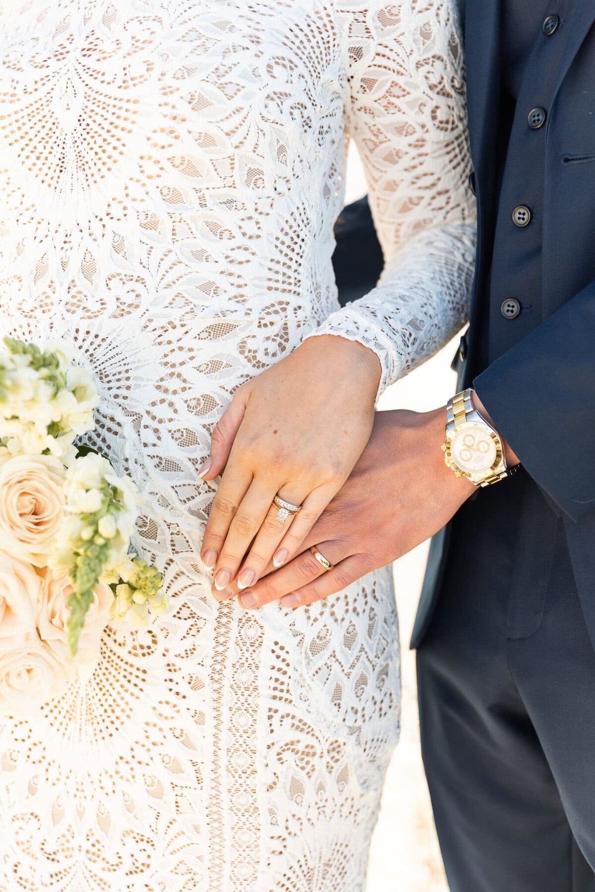 A close-up of the bride’s hand resting gently on her groom’s, their wedding rings gleaming in the soft light. Her intricately patterned lace sleeve adds a touch of timeless romance, while a bouquet of blush roses and white florals peeks into the frame