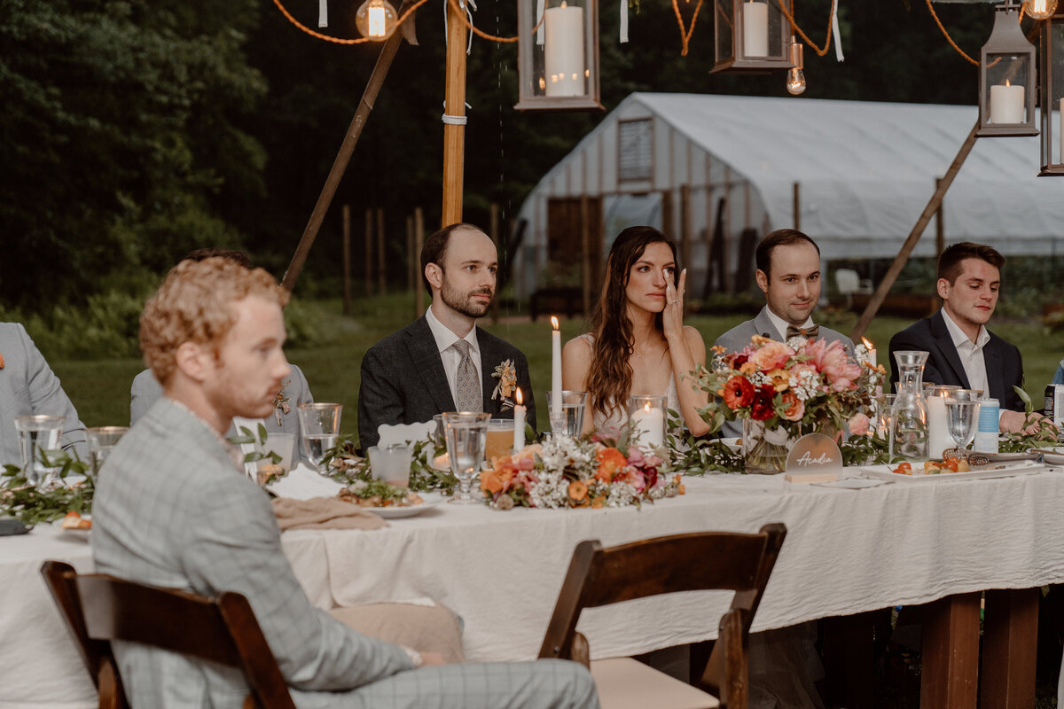 Bride and groom seated at an elegantly decorated table during an outdoor reception, featuring floral arrangements, candles, and lanterns. Bride away a tear, capturing the emotional and heartfelt atmosphere of the celebration. The rustic setting includes a greenhouse in the background, enhancing the natural and intimate feel of the event.