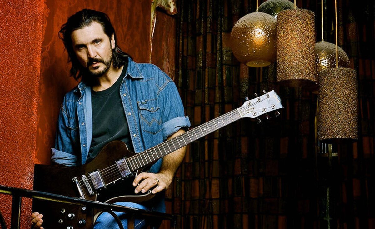 Gordie Johnson portrait wearing blue denim shirt holding dark wood electric guitar leaning against railing large golden lamps behind