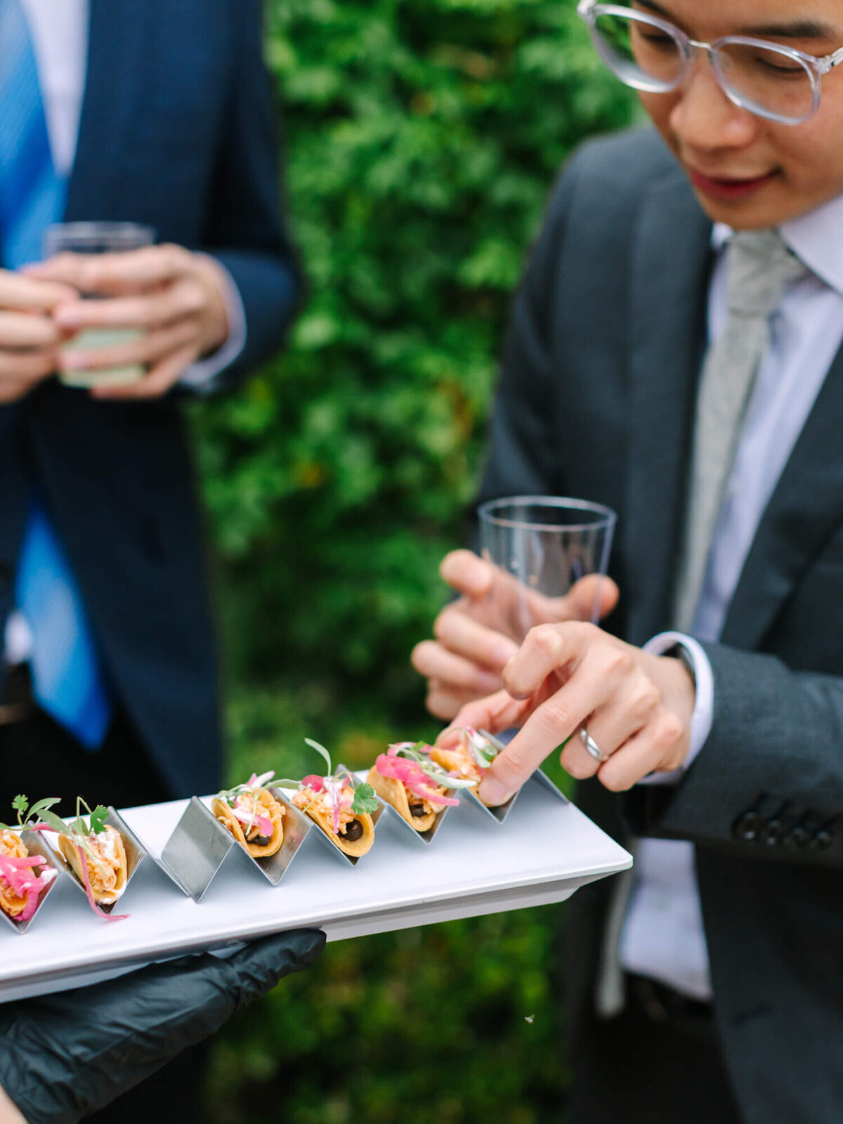 A person in a suit reaches for mini tacos on a tray held by a gloved server. Another person holds a drink. The setting is a formal outdoor event.