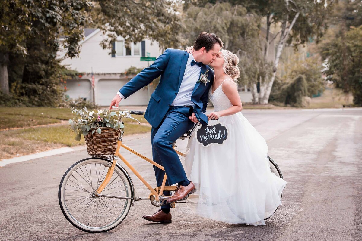 Bride and groom kissing while on a yellow bicycle with a basket of flowers holding a sign that says just married