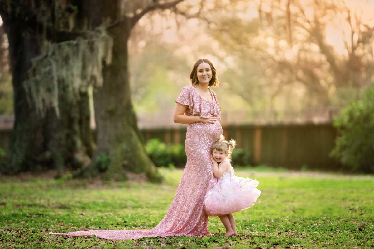 Pregnant mom in a pink sequin Mii-Estillo dress outdoors in Audubon Park.  The woman has short brown hair and is cuddling her 3 year old daughter, who is wearing a white and pink floral Anna Triant Couture Dress.