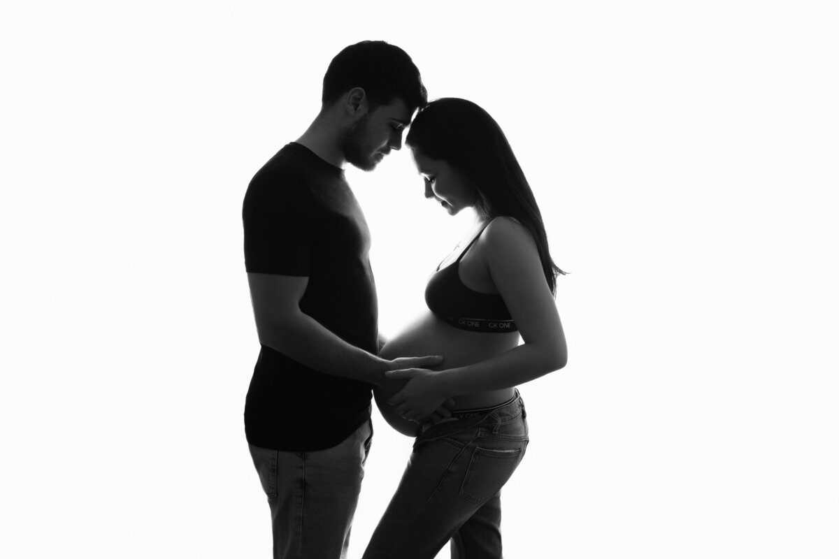 Silhouette of a pregnant couple lovingly facing each other with both of their hands on her belly.