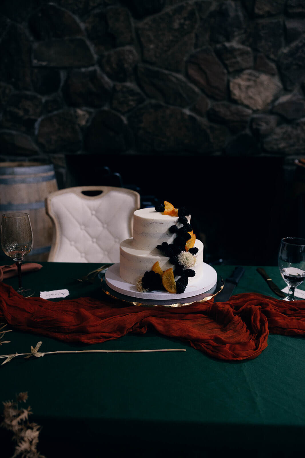 A white two tiered wedding cake sitting on a table and decorated with fruit and flowers.