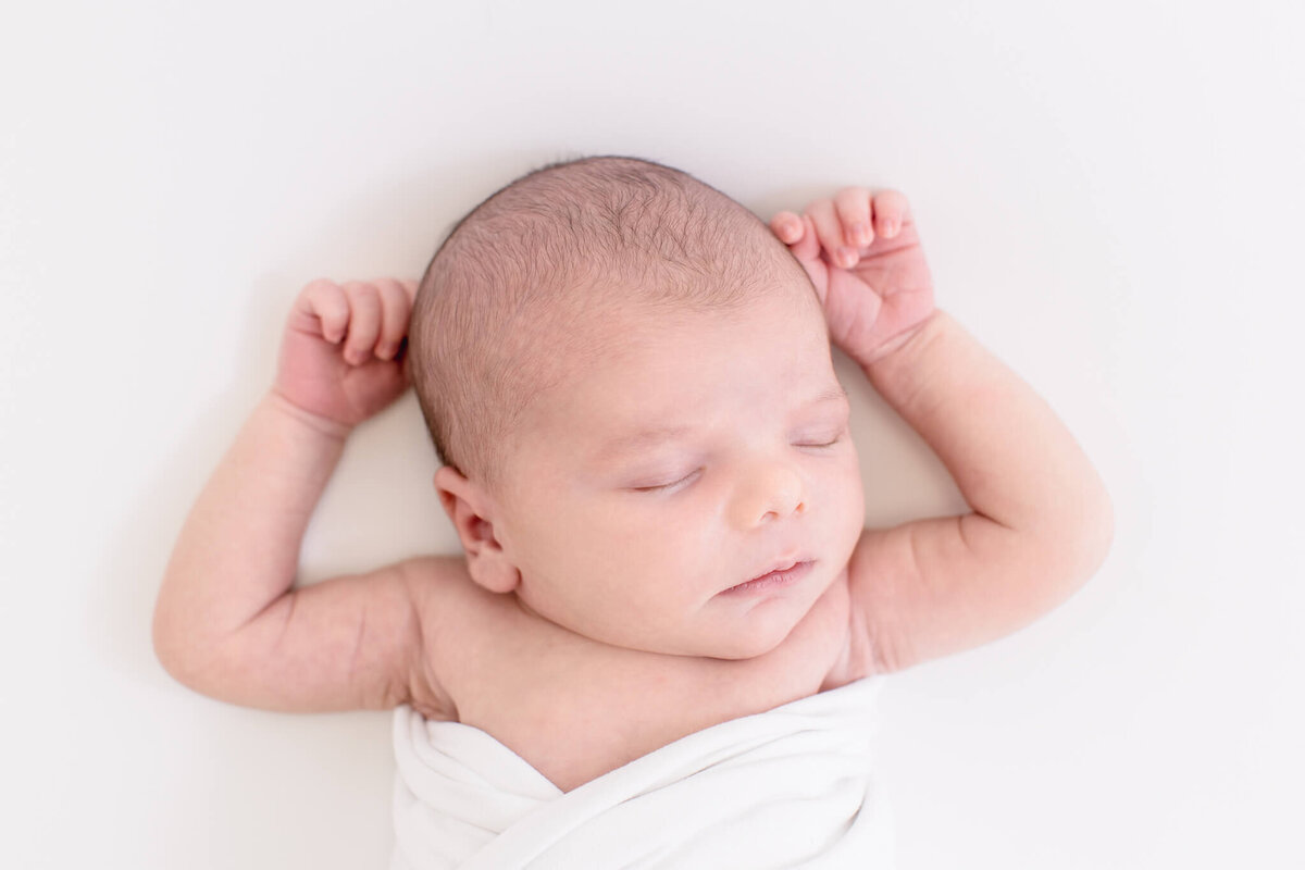 A peaceful newborn baby is sleeping on a white background, wrapped in a soft white blanket. The babys arms are gently raised above their head, and they have a calm expression.