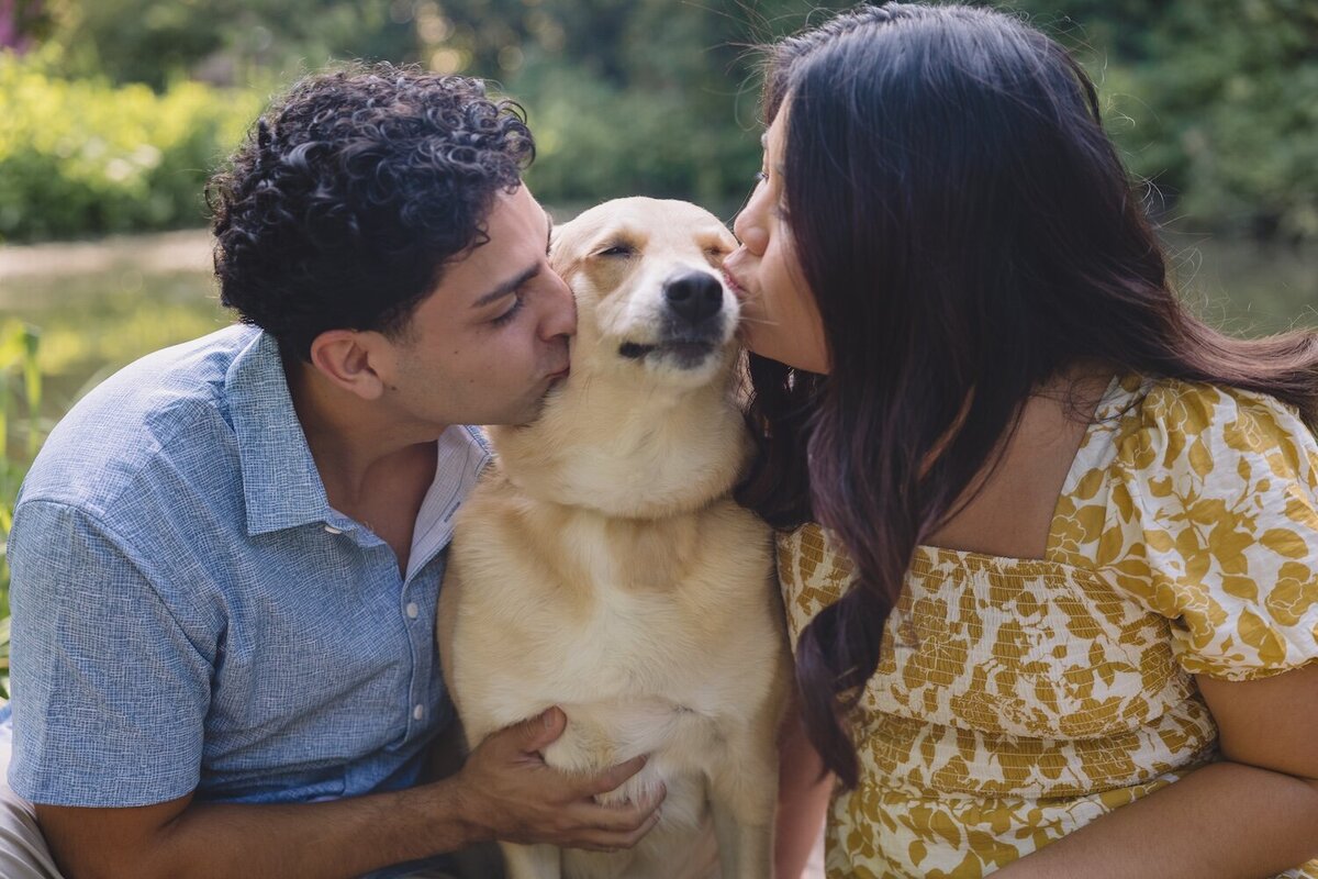 Couple kissing their dog while sitting on a path surrounded by greenery.