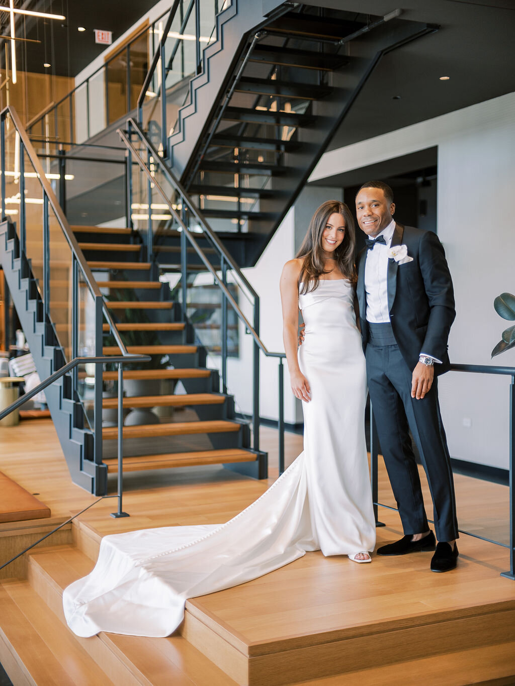 The newlyweds pose in a contemporary space, standing near a sleek black staircase.