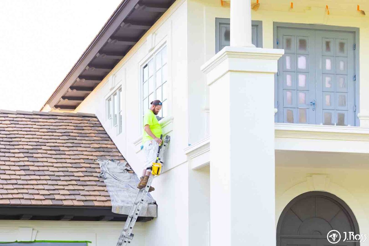 Painter standing on a ladder applying a clean finish to the stucco wall next to a large white column, highlighting the detailed work required for professional home exterior painting Bristol, VA.