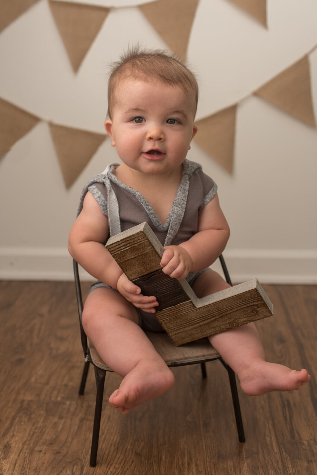 Infant boy sitting on a small metal chair, holding letter L with burlap banner behind him.