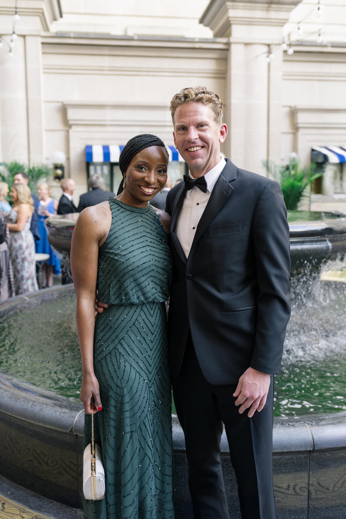 A couple poses in front of a fountain, smiling. The woman wears a dark green sleeveless gown, and the man is in a black tuxedo. They stand closely, with people and greenery visible in the background.