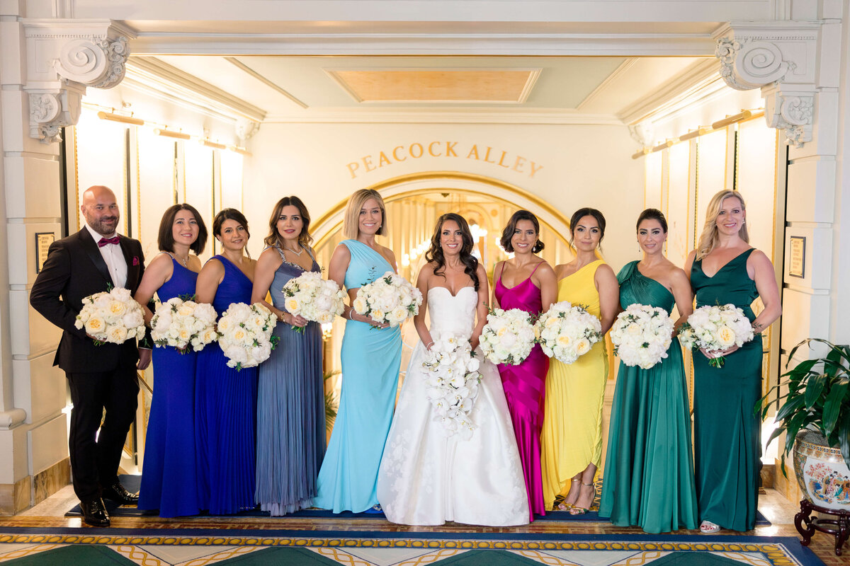 A bride in a white gown stands in a line with a groom and bridesmaids wearing dresses in various colors. They all hold white floral bouquets. The setting is a elegantly decorated corridor with Peacock Alley sign in the background.