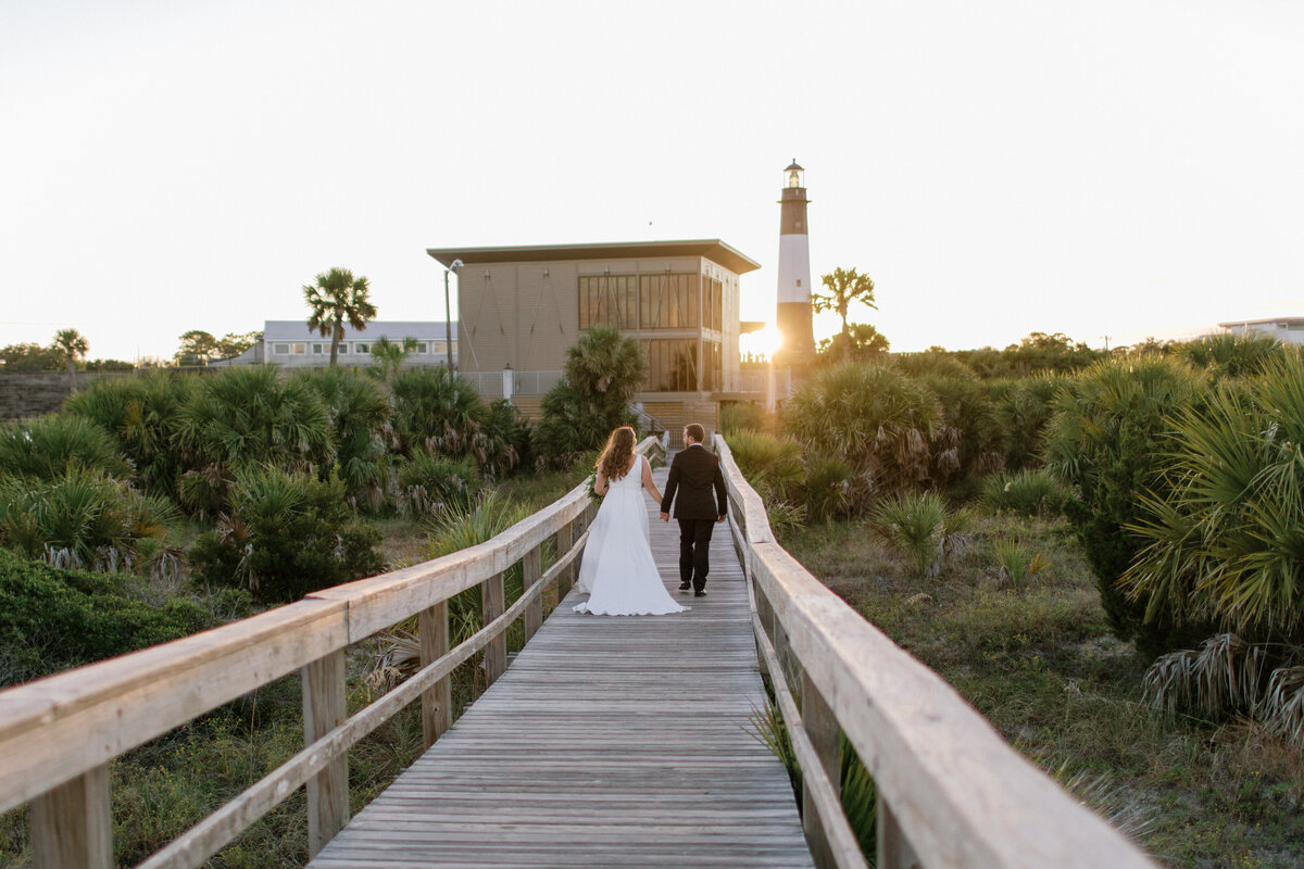 Wedding photography in Statesboro, GA of bride and groom walking away from camera down beach board walk holding hands at sunset toward a lighthouse with sunset peeking around the side.