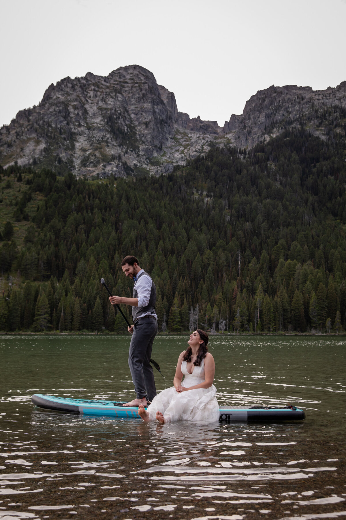 A bride sits on a paddleboard while her groom paddles around a lake in Wyoming.