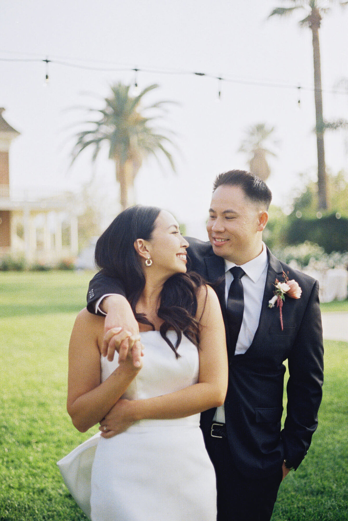 A joyful couple stands on a sunlit lawn, smiling and holding hands.