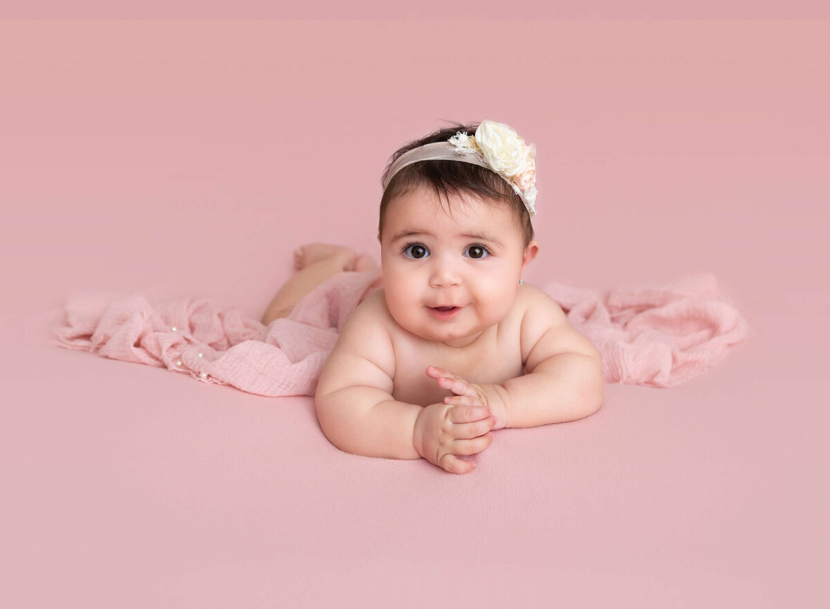 6-month baby girl is lying on her belly for a milestone photoshoot. Baby has her head lifted and is looking at the camera.