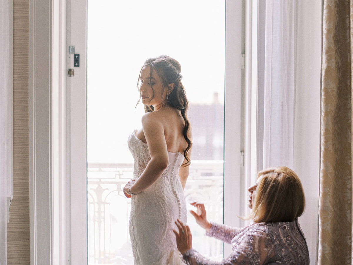 A bride in a white dress stands by a large window, smiling, while another woman adjusts the back of the dress during a classic Calgary wedding.