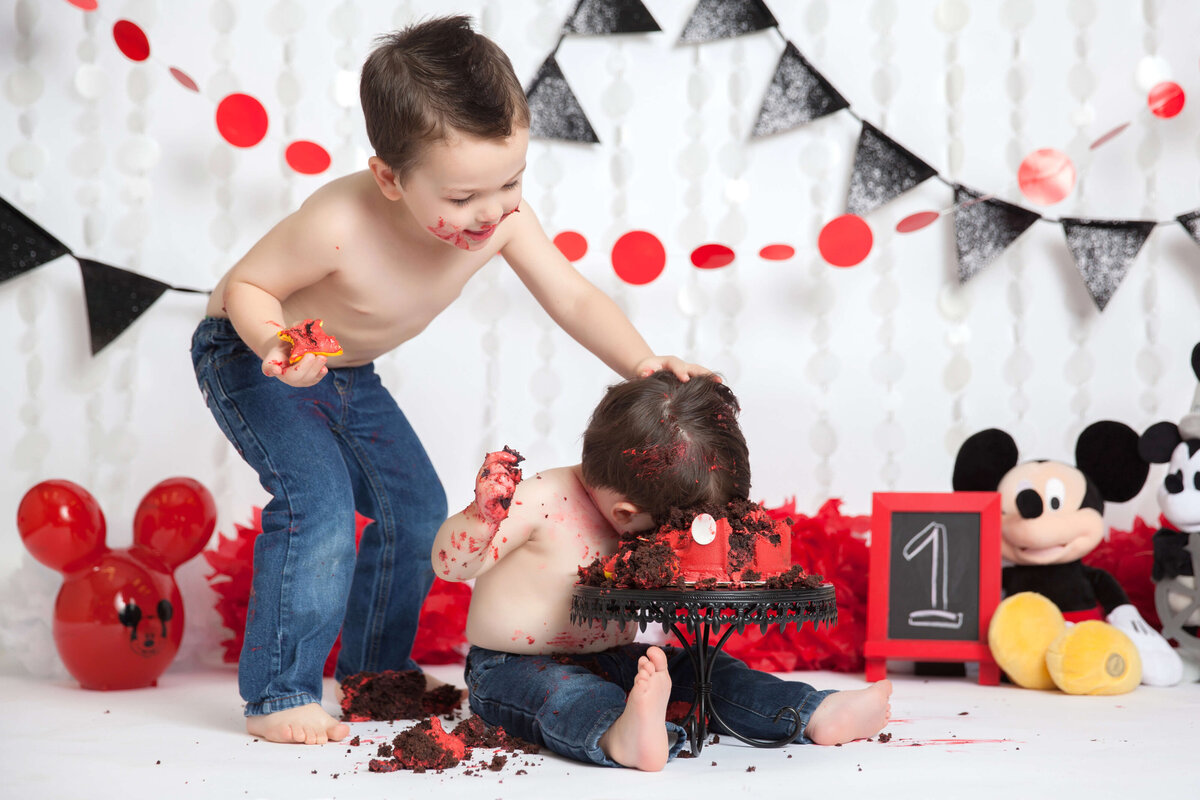 Two young children in jeans are celebrating a first birthday with a delightful cake smash. One child playfully smushes cake onto the seated other's face. Red and black banners, a chalkboard with "1," and Mickey Mouse toys set the scene, captured beautifully by a Jacksonville cake smash photographer.