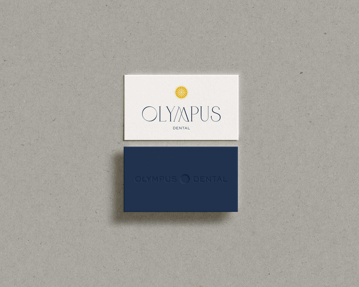 Two business cards for Olympus Dental. The top card is white with black text and a yellow sun logo, showcasing their brand designer's flair. The bottom card is dark blue with subtle text and logo, difficult to read. Both are on a gray surface, reflecting thoughtful brand strategy consulting.