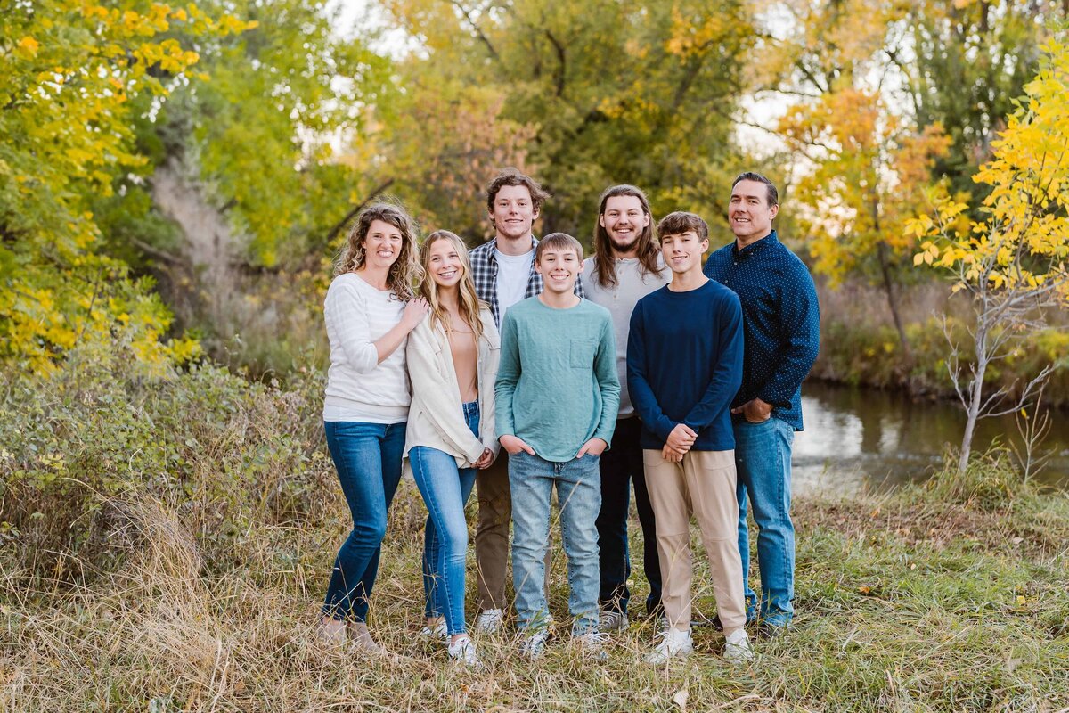 Family of seven standing together near a creek surrounded by fall colors, smiling warmly during a relaxed outdoor portrait session by a Colorado Wedding and Portrait Photographer.