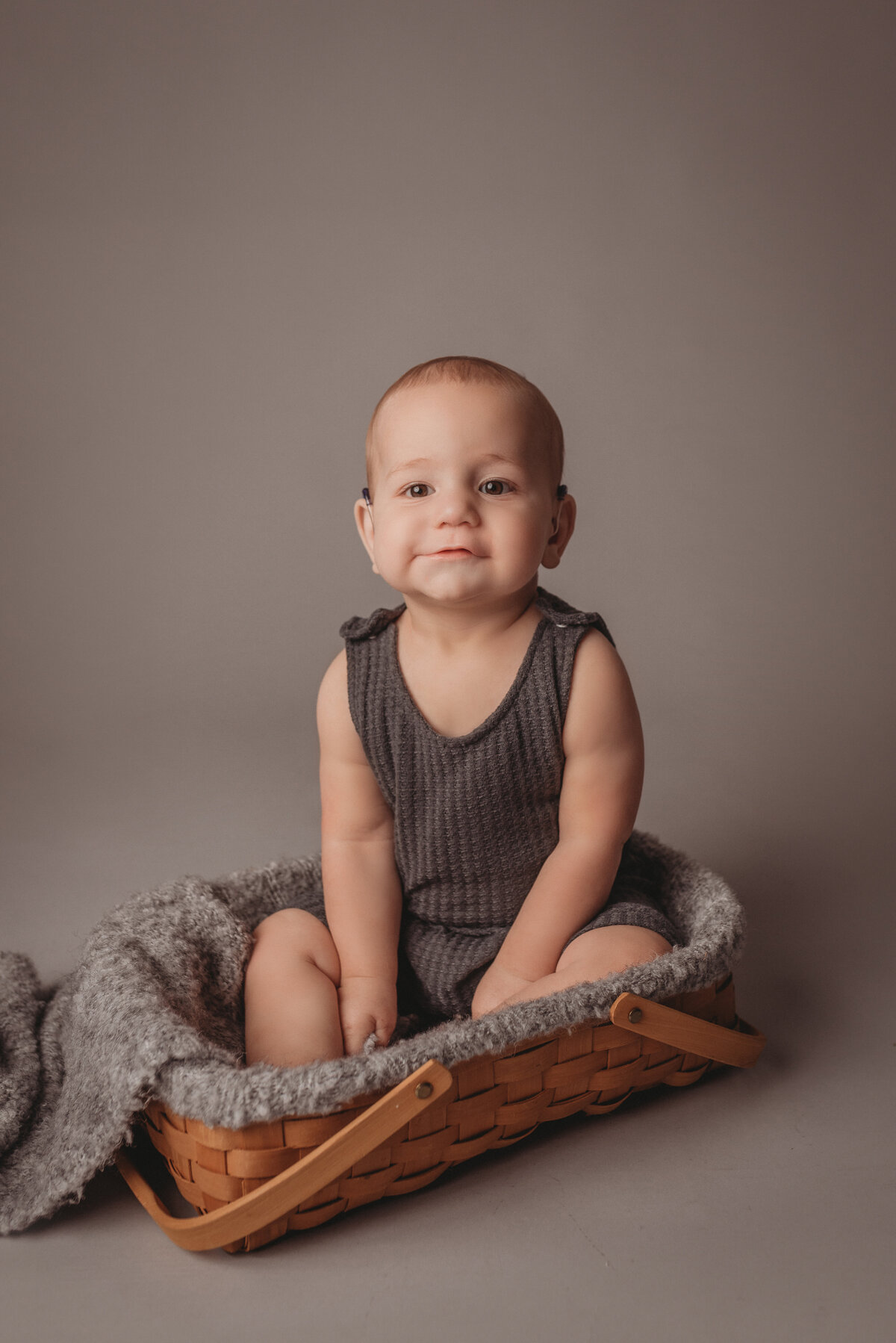 One year old baby boy wearing gray waffle knit romper sitting in basket posing for his portraits on a light gray backdrop