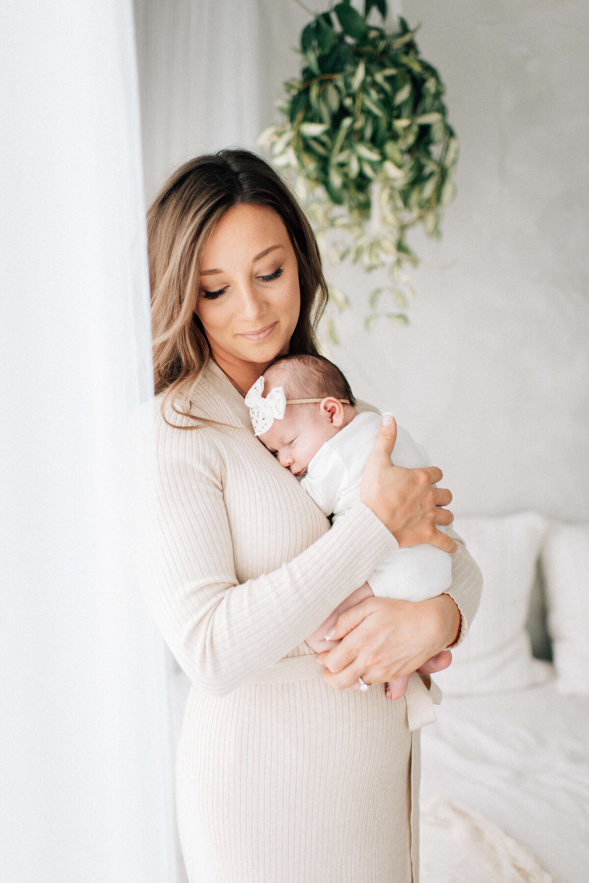 mother holding baby girl close to her chest near a window with a plant hanging in background