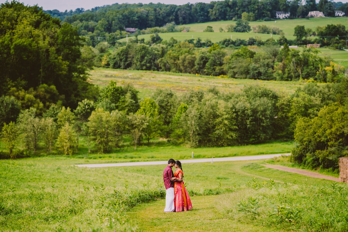A wide shot on top of a hill capturing an engaged couple embracing down the hill with the natural lands stroud preserve in the background