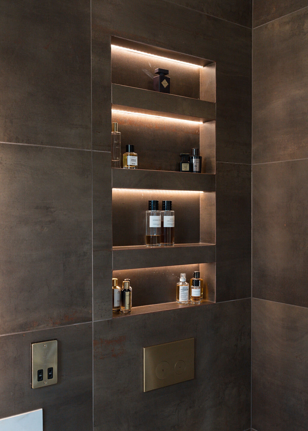 Above a toilet in a modern bathroom, lit shelves are built into the wall, holding toiletries and emitting a soft light. The wall has large, dark brown tiles.