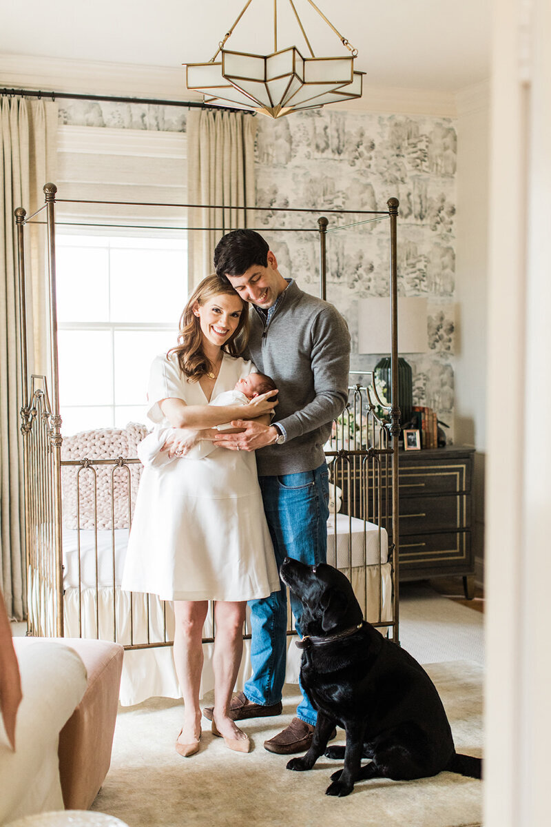 parents stand holding newborn in nursery near crib with their black lab