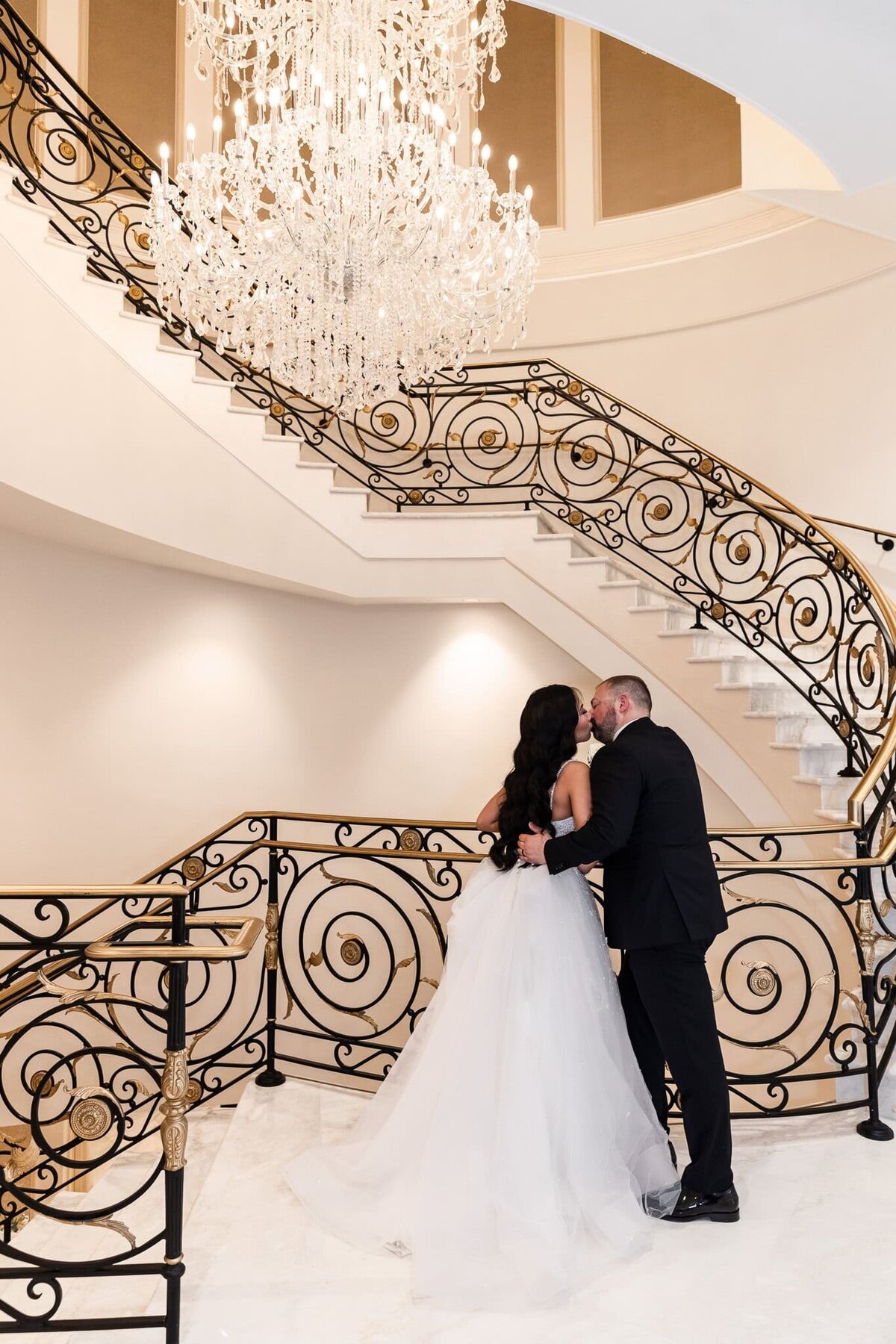 A newlywed couple shares a quiet kiss at the base of a grand, sweeping staircase adorned with intricate gold and black wrought iron railings. A massive crystal chandelier illuminates the moment, casting a soft glow on the bride’s voluminous tulle gown and the groom’s classic black suit