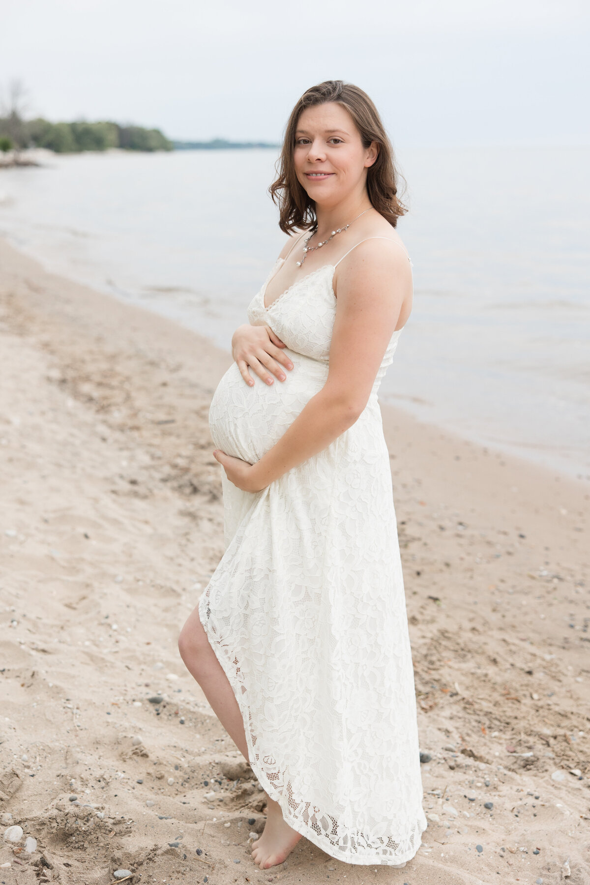 Expectant mother holder her baby bump on the beach and smiling at the camera