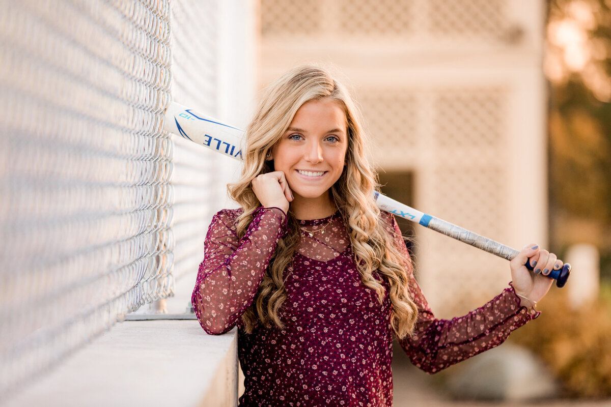 A young woman smiles with her baseball bat near a fence.