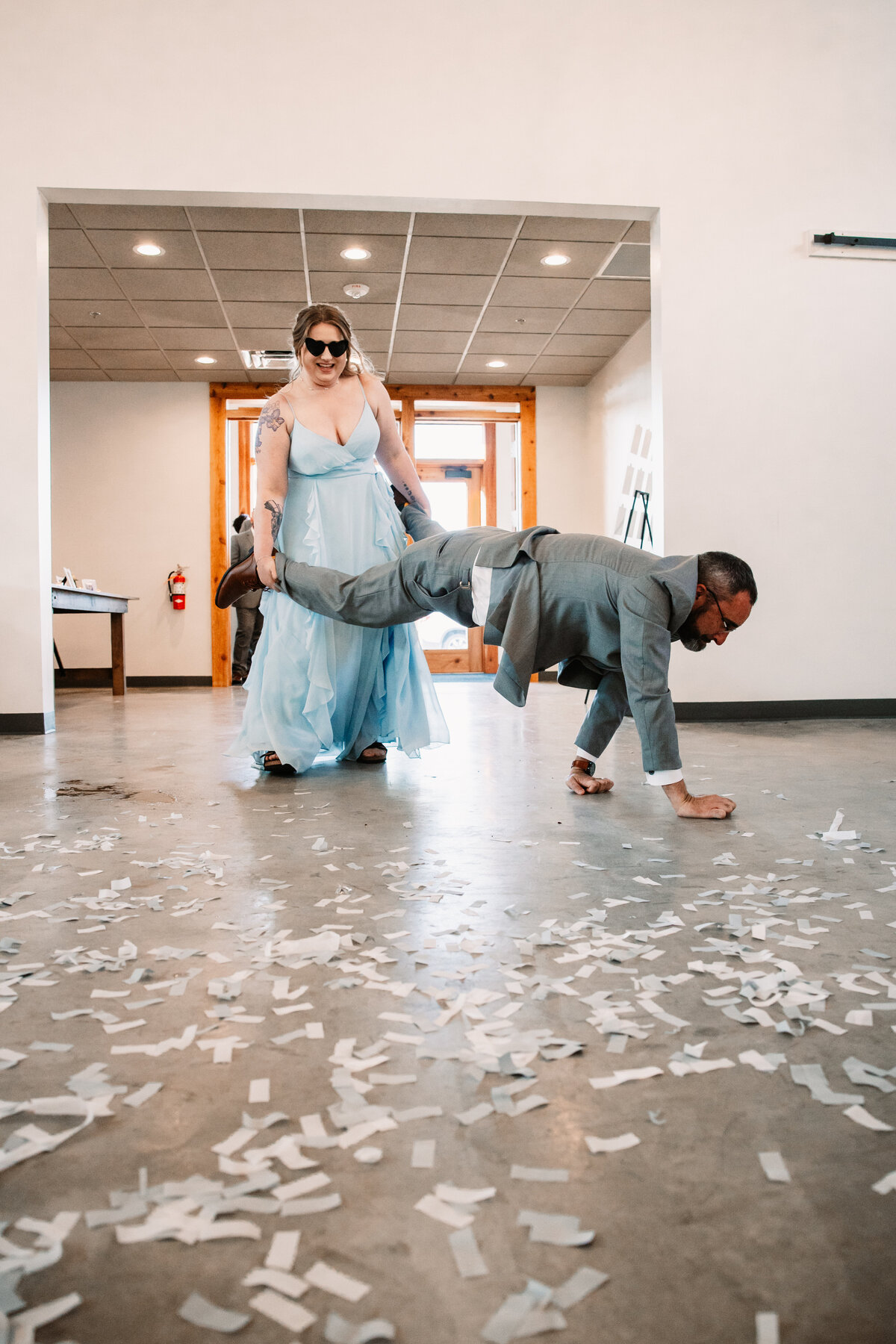 A bridesmaid enters the reception holding the legs of a groomsman as he walks on his hands with confetti everywhere