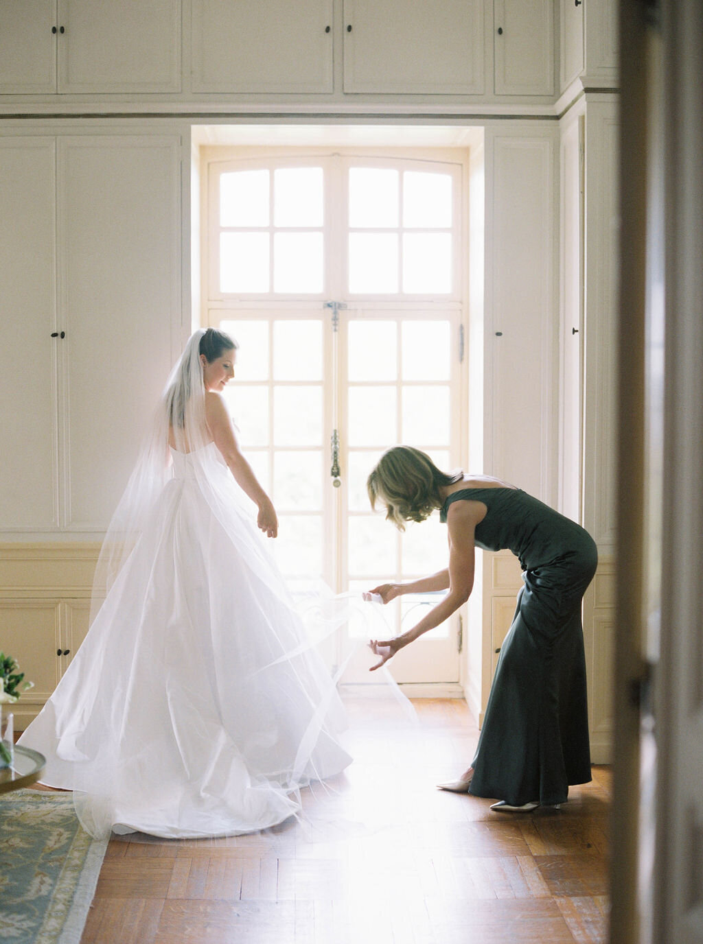 Glen Manor House | Bride in flowing gown stands by a bright window, assisted by a woman adjusting her train. 