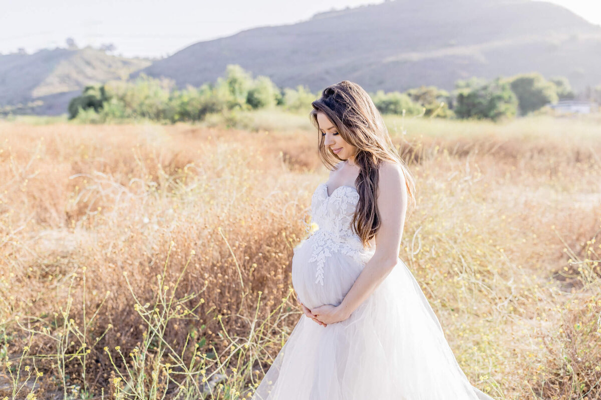 A pregnant woman in a white dress stands in a field, gently cradling her belly. The background features hills and trees under a clear sky, creating a serene and natural setting.
