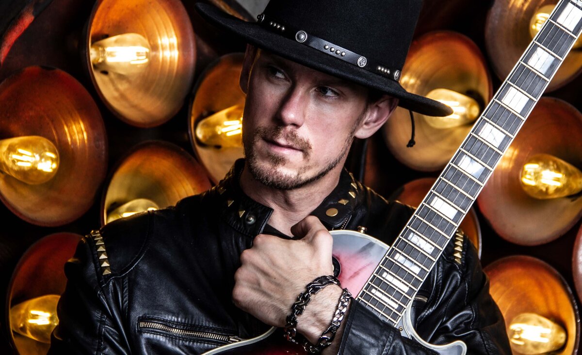 Male musician photo wearing black leather jacket black cowboy hat clenching red electric guitar gold circle lights behind
