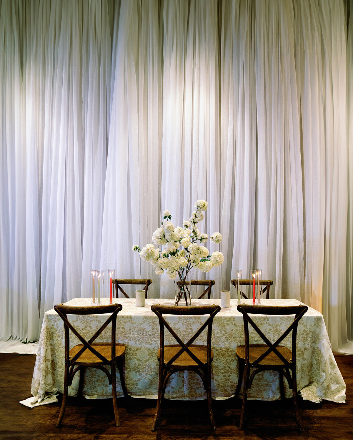 Elegant wedding reception table at Burritt on the Mountain with white floral centerpiece, patterned tablecloth, wooden cross-back chairs, and tall draped backdrop.