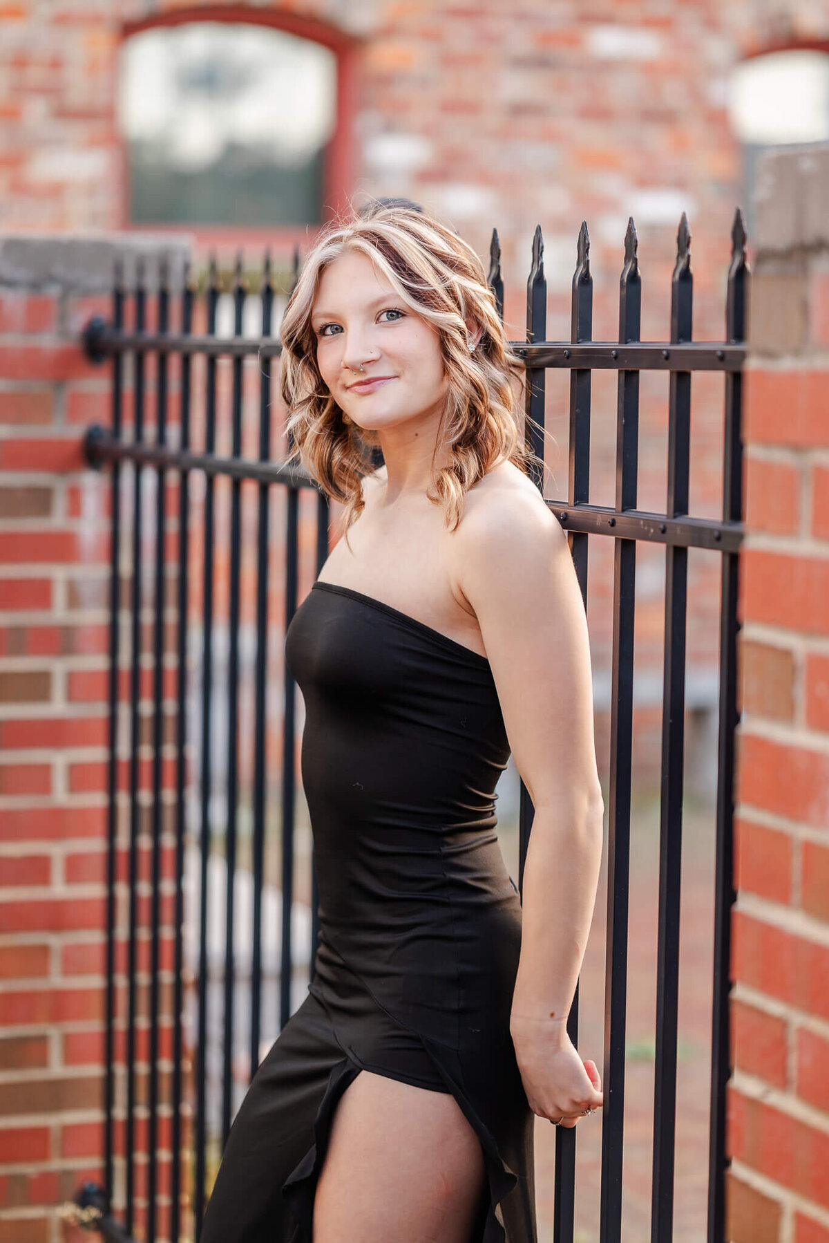 A high school senior, wearing a black dress with a slit, leans on a wire fence in a brick wall. She is having her senior session with Chesapeake senior photographer, Justine Renee Photography.