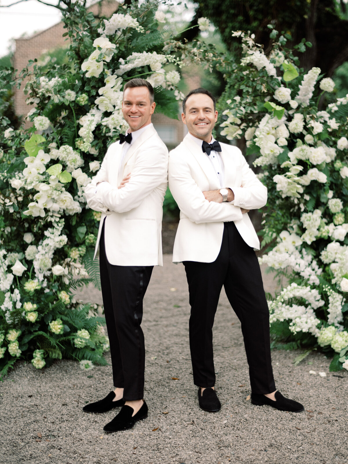 A charismatic couple in white tuxedos poses confidently outside, surrounded by beautiful floral arrangements, showcasing love and unity during their special day.