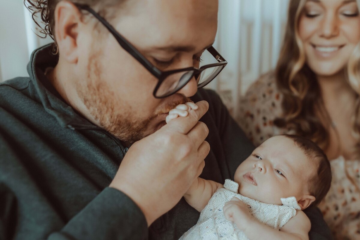 A man wearing glasses gently kisses a baby's hand while a woman smiles in the background. The baby is wearing a white knitted outfit.