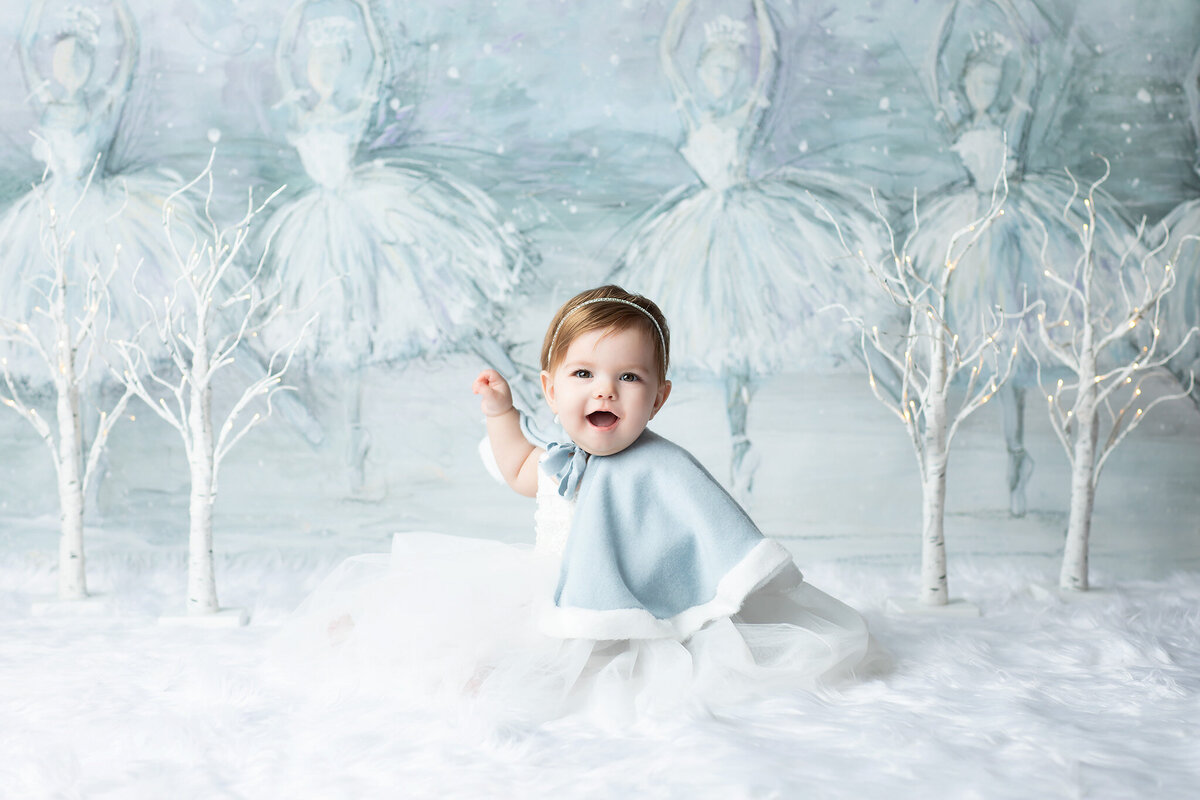 Baby girl dressed in a soft blue cape and white tulle skirt sits on faux snow in front of a dreamy ballerina backdrop, styled by a Dallas milestone photographer.