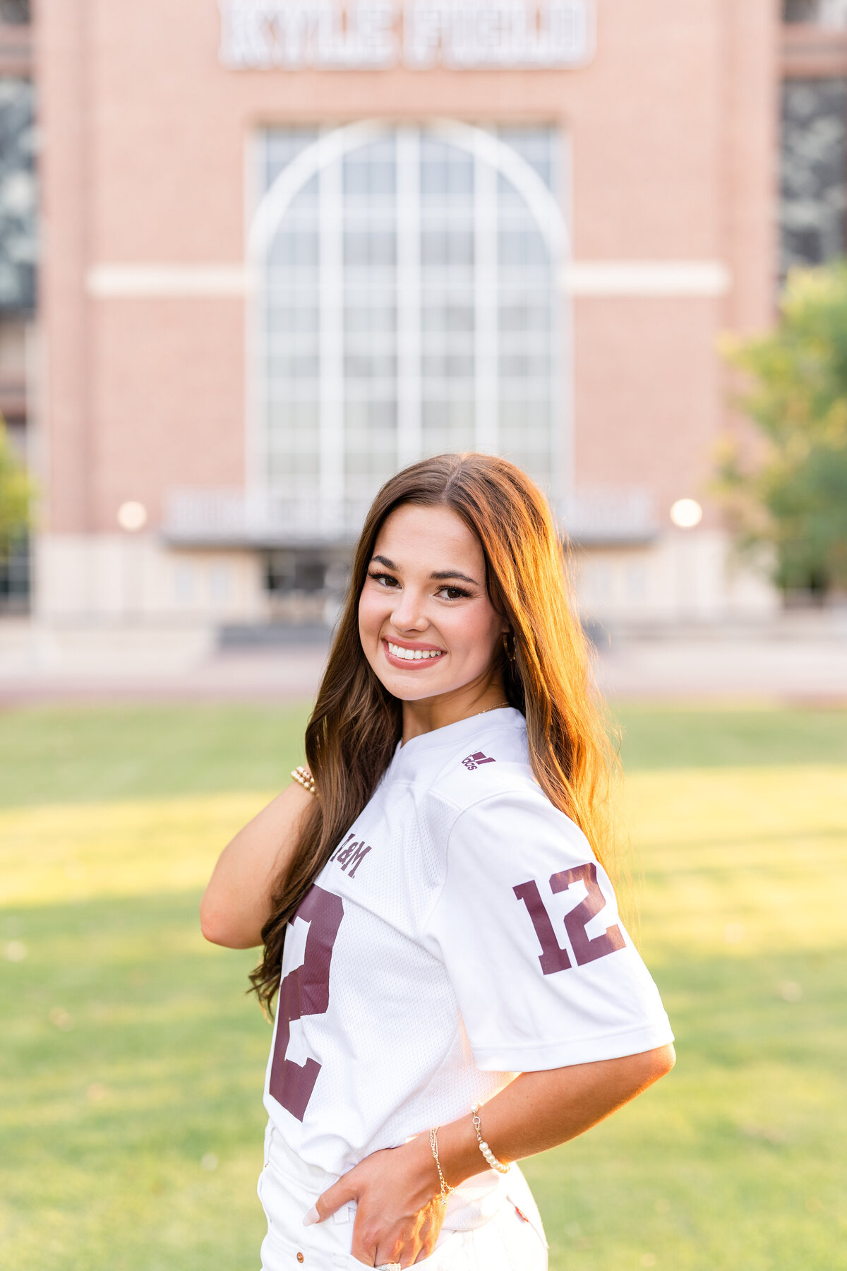 Texas A&M senior girl wearing white Aggie jersey with hand in back pocket and other hand in hair while looking back over shoulder and Kyle Field in background in Aggie Park