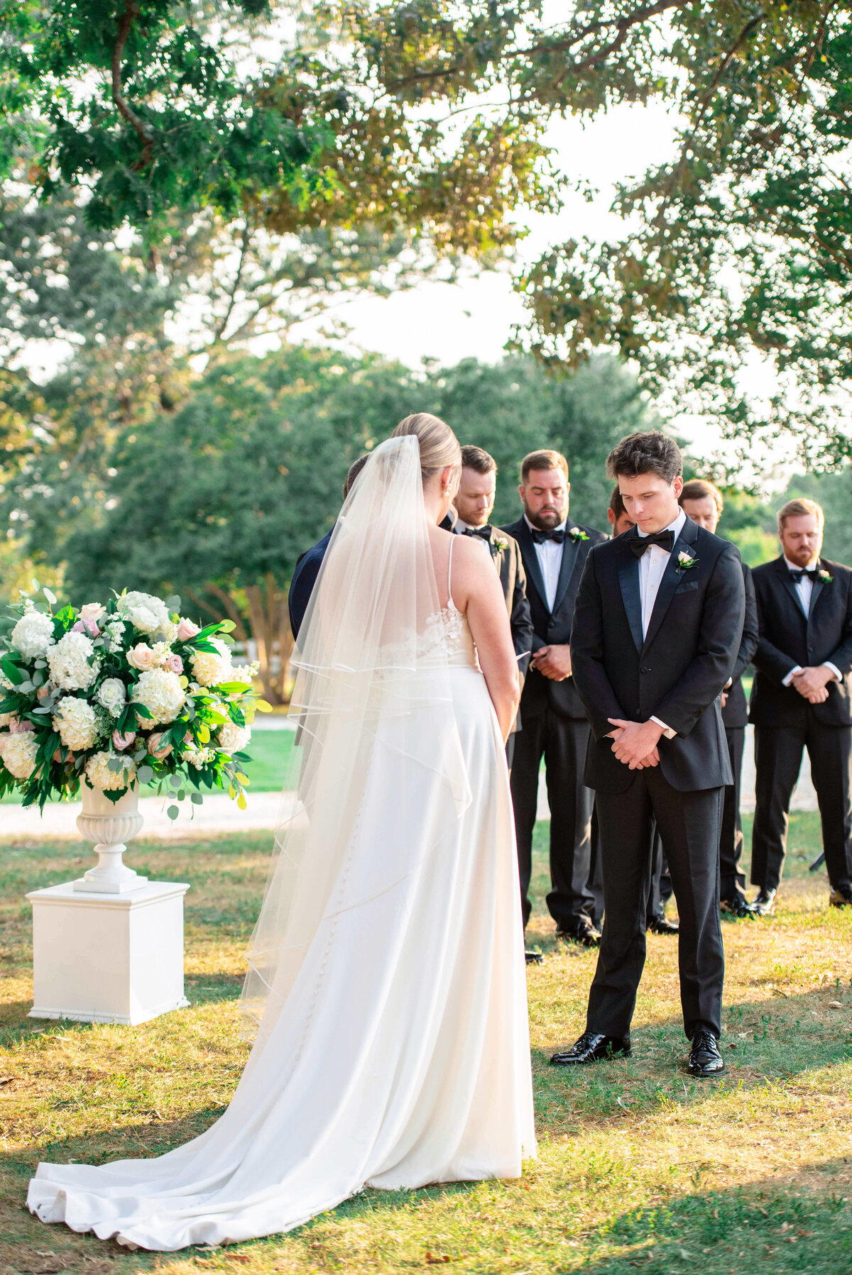 A bride and groom stand facing each other during an outdoor wedding ceremony. The bride wears a white gown and veil, while the groom is in a black tuxedo. Groomsmen stand nearby, and a large floral arrangement is visible. Trees are in the background.