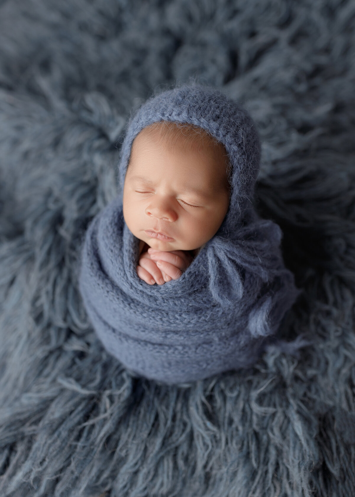 Newborn photoshoot with Lake Worth newborn photographer. Baby is wrapped in a blue knit swaddle with his hands peeking out of the top. Baby boy is sleeping wearing a matching blue knit bonnet. Baby is sitting atop of a matching blue furry rug.