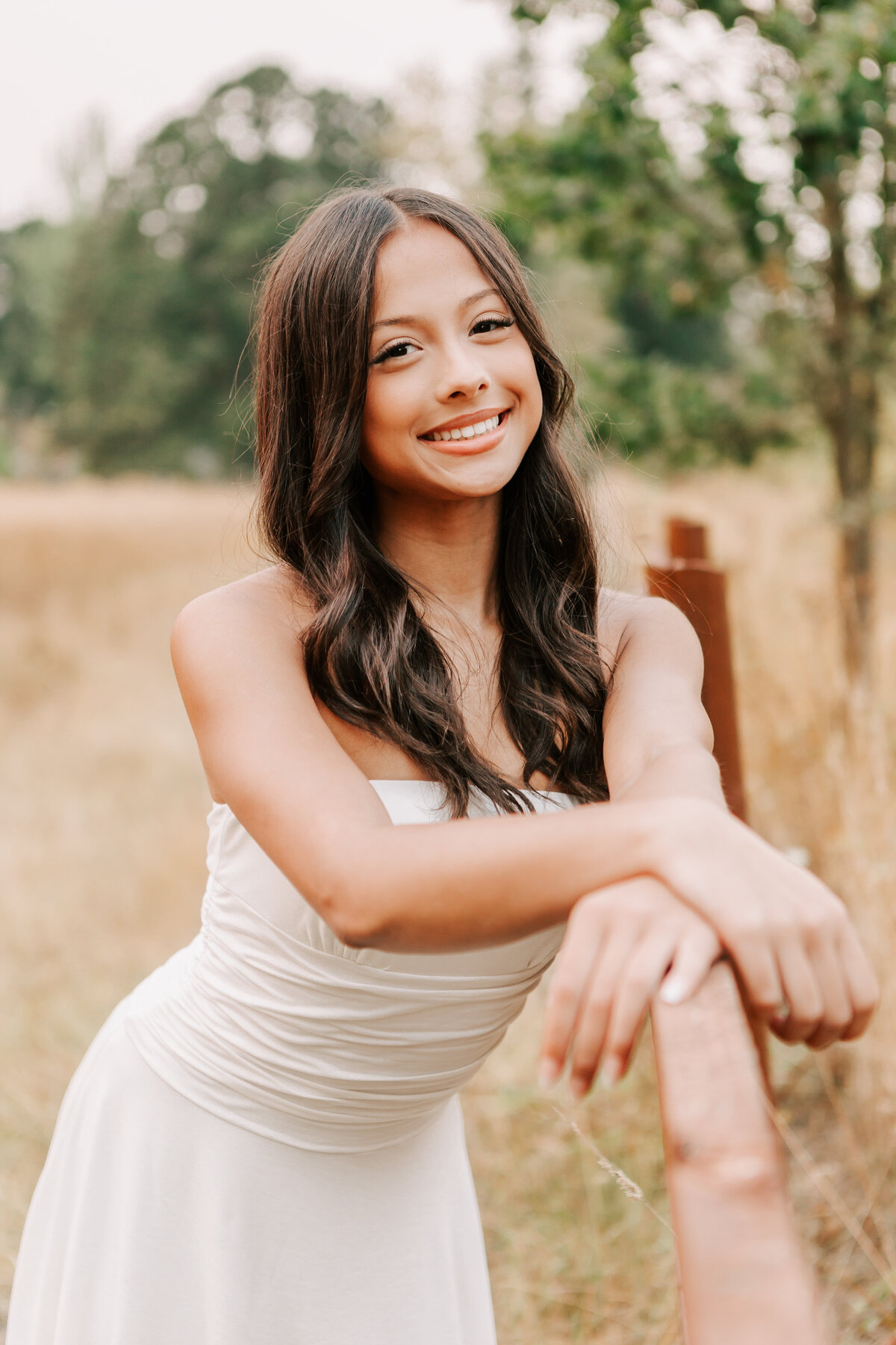 A Senior girl in a white dress leans on a fence at a farm in Albany, OR